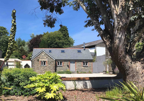 Between shrubs and a mature tree, stands a Cornish 2-storey stone-barn conversion