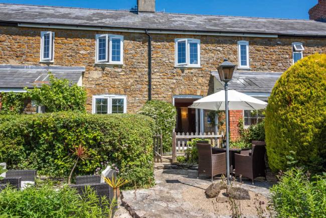 Exterior and front garden of a stone-built Dorset holiday cottage with outdoor dining furniture on its terrace.
