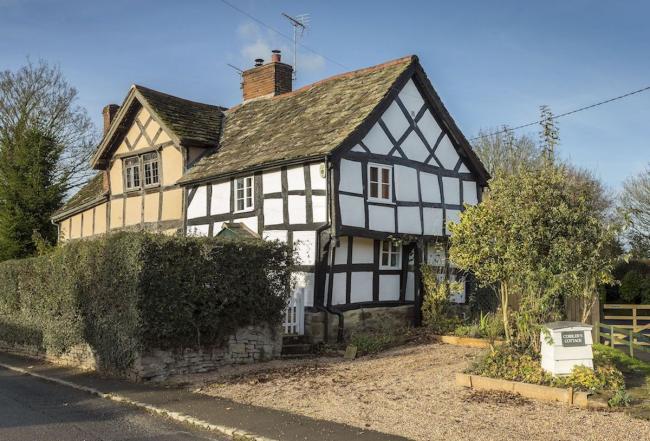 A half-timbered Tudor cottage  in Leominster overlooks a shingle parking space and shrub-filled garden.