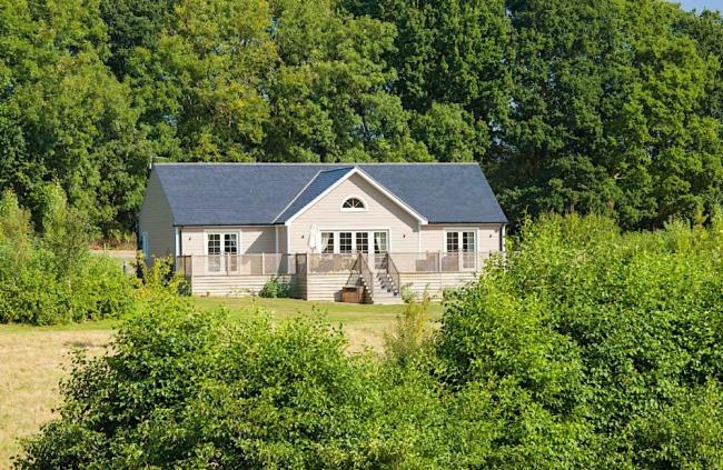 A modern single storey lodge stands in front of trees overlooking a hedge-lined field.