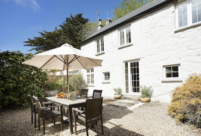 A stone-built cottage overlooks a shrub-lined terrace with dining table chairs and a parasol