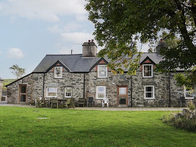A wide, 2-storey, stone house in Snowdonia with three gable ends flanked by single storey wings facing a large, level lawn with mature trees.