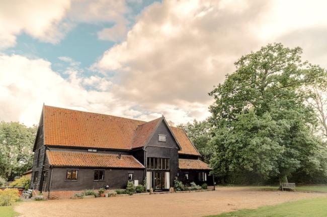 Exterior of a large Manor Farm Barn made of wood surrounded by a tree-filled garden.