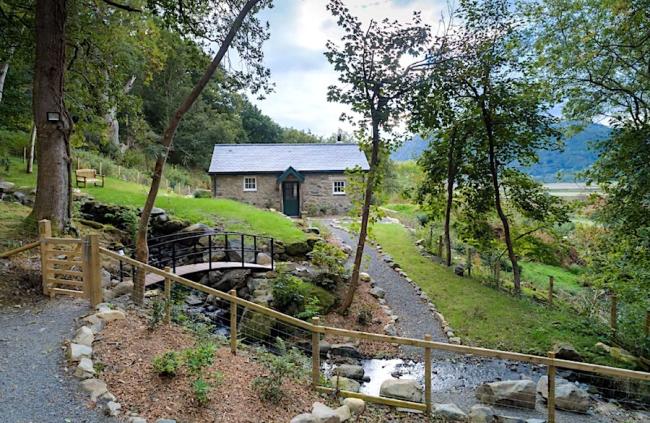 A single storey, stone cottage overlooks a garden with a small stream trickling under a bridge in the foreground.