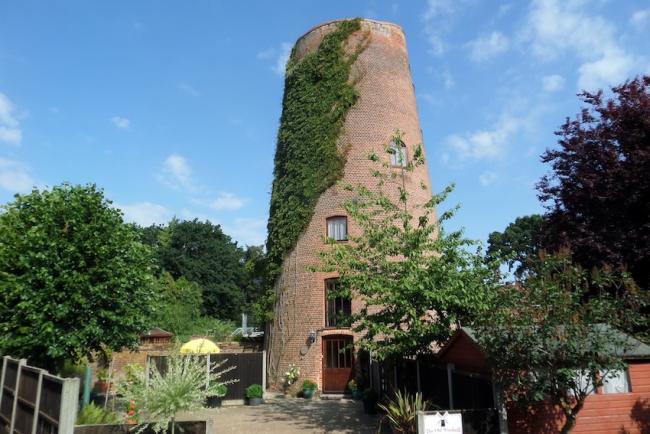 The tower of a windmill (no sails) surrounded by a shrub-filled garden.
