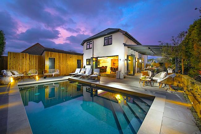 A large, brightly lit house overlooks a swimming pool at dusk.