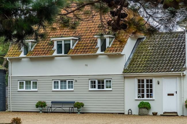 Exterior of a weather-boarded Suffolk Cottage behind a garden lined with fur trees.