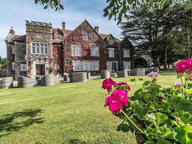 Frontage of a large 3-storey, Devon country house overlooking an outdoor pool and tree-lined lawns.
