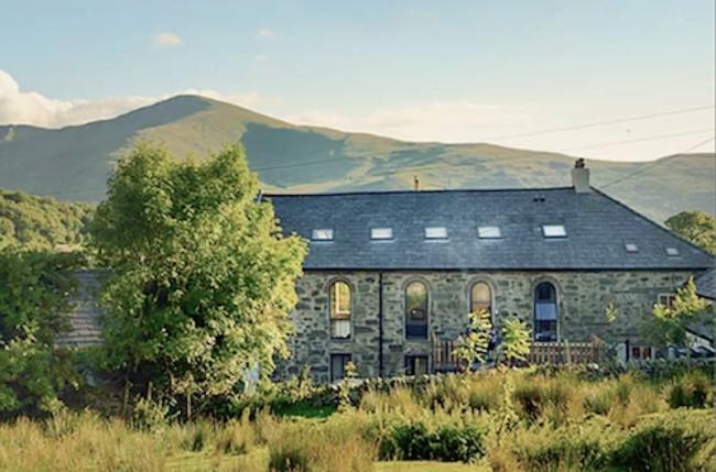 A stone barn conversion backed by mountains overlooks a field with small trees.