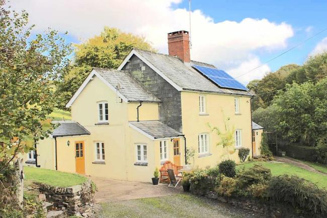 A 2-storey-cottage overlooks a lawn surrounded by trees.
