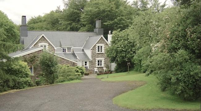 A sweeping drive approaches a twin-gabled Exmoor holiday cottage half-surrounded by mature trees.