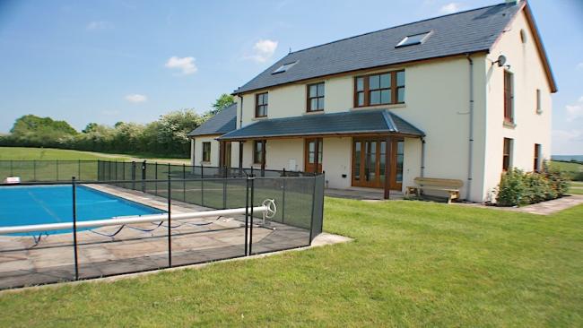 A fenced-in outdoor pool on a lawn overlooked by a large holiday cottage.