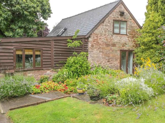 The exterior of a stone-built cottage with a wooden extension overlooks a flower-filled garden.