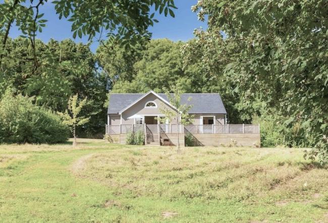 On the edge of a mature wood, a timber-frame lodge with a spacious deck overlooks an open field.