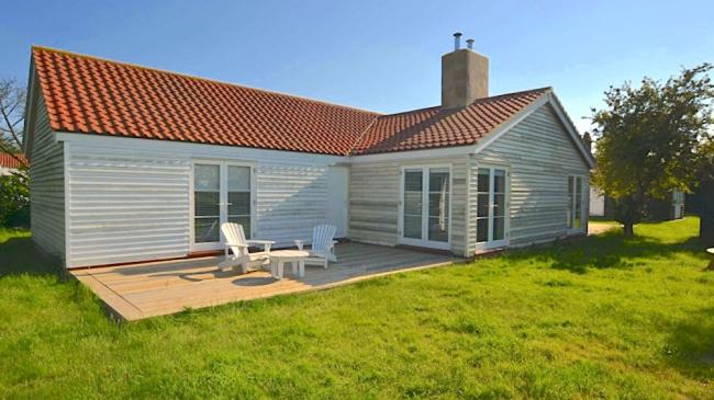 An L-shaped, wood-panelled Essex holiday bungalow with a wooden deck containing a table and sunlounger, all overlooking a large lawn.