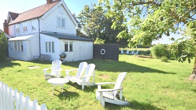 A wooden 2-storey Essex holiday cottage overlooks a lawned garden, shaded by tall trees with four sun loungers on the grass.