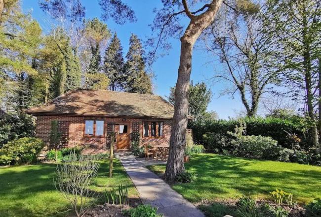 The exterior of a brick-built Dorset bungalow is surrounded by lawns, shrubs, and mature trees.