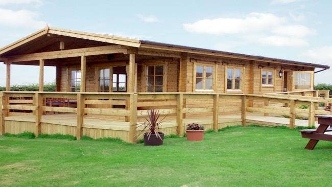 A long eco-friendly log cabin with a covered, wrap-around deck is surrounded by countryside..