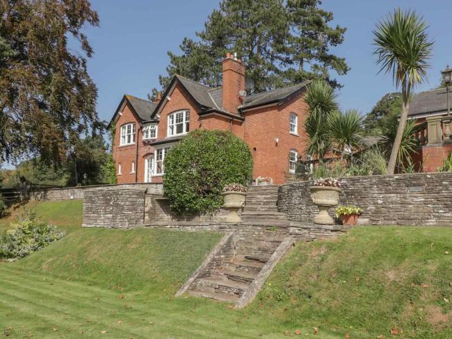 A large brick house backed by tall pine trees overlooks an extensive lawn.