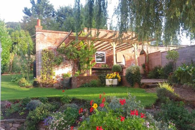 The exterior of a single-storey holiday apartment with a vine-covered pergola and floral garden.