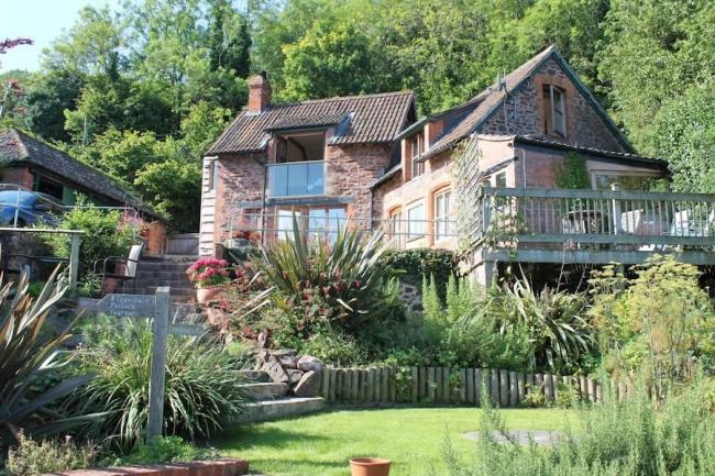 Against a woodland backdrop, a large cottage with a spacious deck overlooks a flower and shrub-filled garden surrounding a lawn.