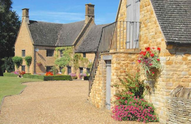Exterior of a flower-decked Cotswold Manor and adjoining barn facing a shingle drive and lawn.