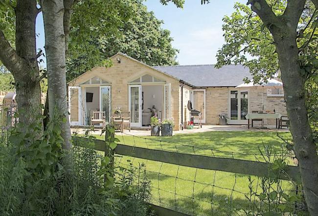 A bungalow with double French Windows onto a patio and a lawn ringed by trees.
