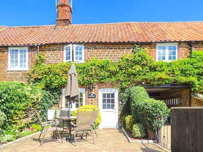 Exterior of a wisteria-clad cottage-front facing a paved front-garden with outdoor dining table, chairs and parasol.