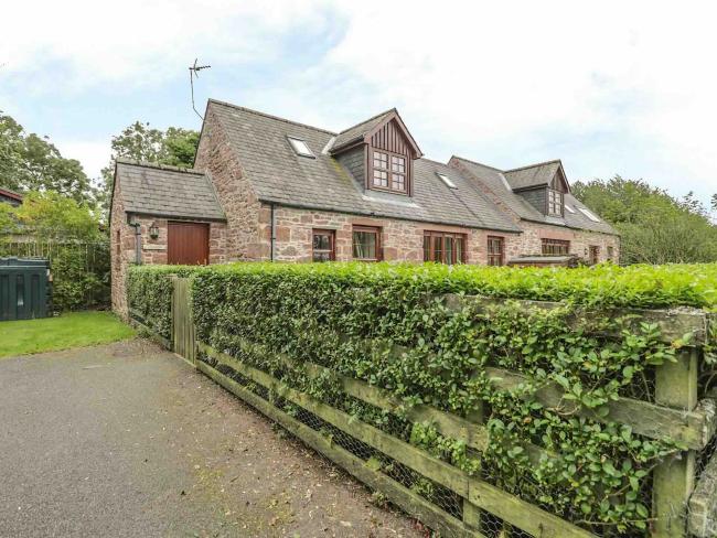 Exterior of a stone-built, semi-detached cottage with a  large, securely hedged-iin garden.