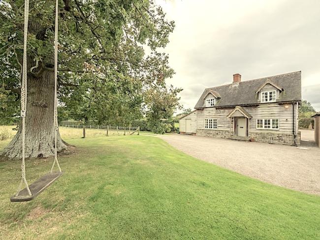 A garden with a garden swing, trees and a wide drive are overlooked by a weather-boarded 2-storey Herefordshire cottage.
