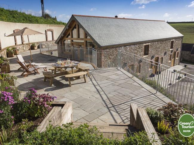 Larger raised and paved terrace with picnic tables and railings overlooks a stone barn conversion and fields.