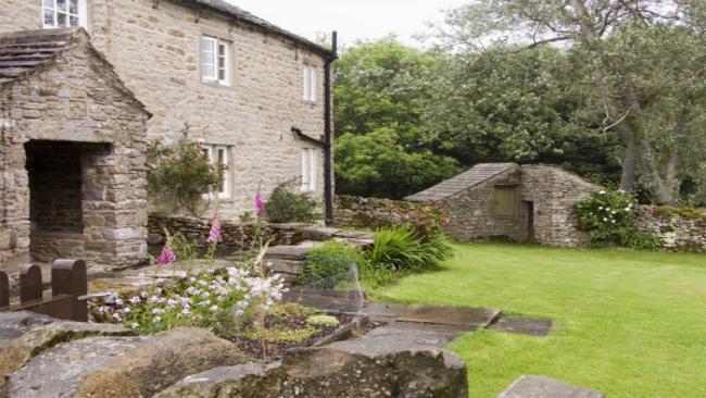 A stone cottage with a large porch overlooks a walled lawn and flower beds.