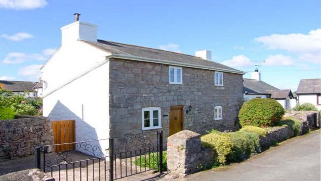 A double-fronted stone cottage  with a hedged-in lawn to the front and a gated driveway for cars.