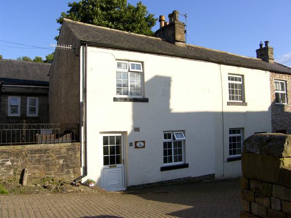 A white-rendered 2-tory, semidetached holiday cottage in Cumbbria overlooks a quiet lane.