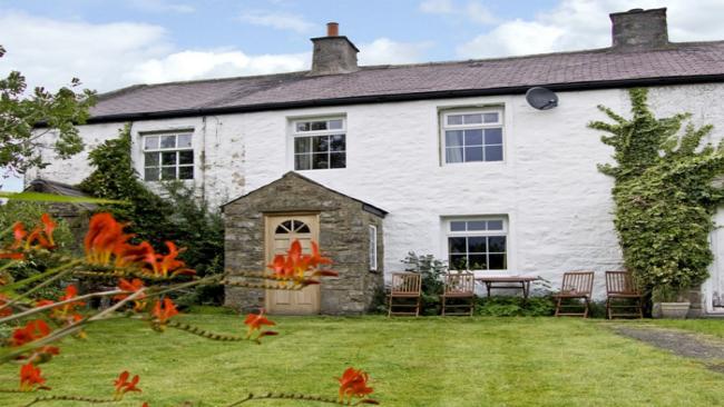 A whitewashed cottage with a stone-built porch overlooks a lawn lined by mature shrubs and flowers.