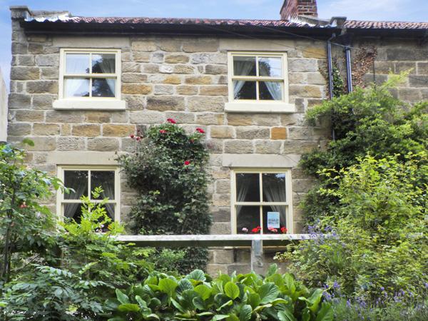 A semi-dtached, stone-buit North Yorkshire holiday cottage sits behind afront garden with shrubs and roses.