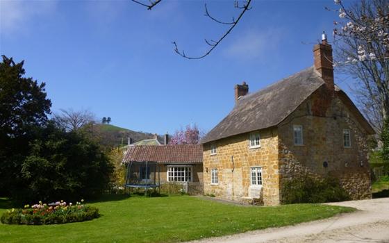 Exterior of a stone-built thatched village cottage overlooking a large lawn with a flowerbed.