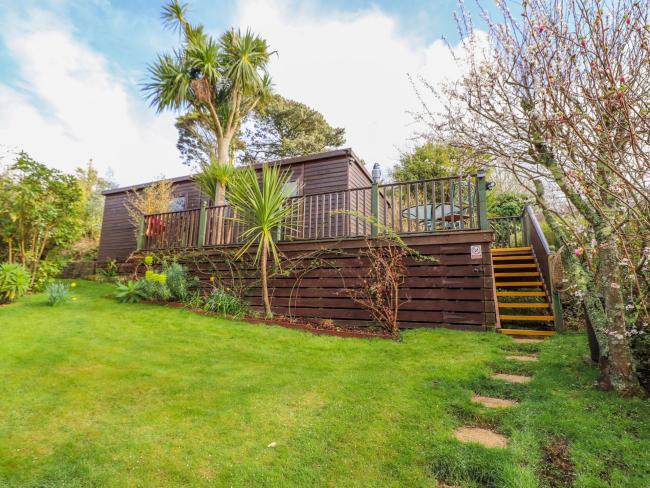 A single-storey timber lodge sits above a raised deck above a large, shrub-lined lawn