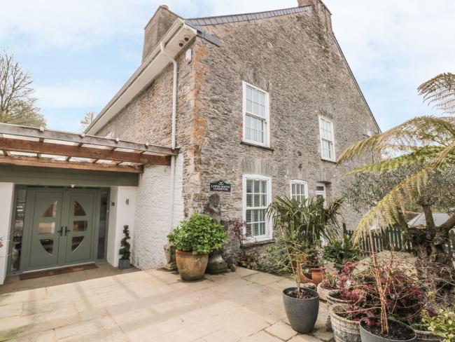 A tall, 2-storey, stone-built cottage overlooking a mature garden with a terrace and palms.
