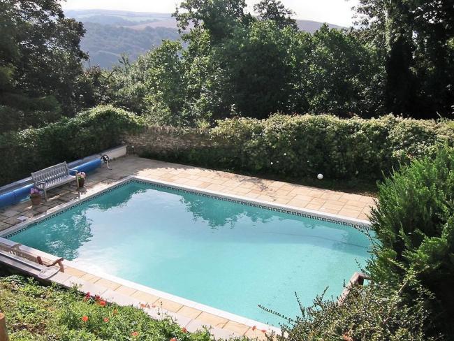 An open air swimming pool with park benches on its terrace surrounded by a tall hedge.