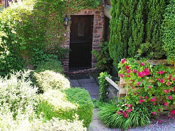 A stout wooden door in an ivy-clad stone-built porch surrounded by shrubs and flowers.