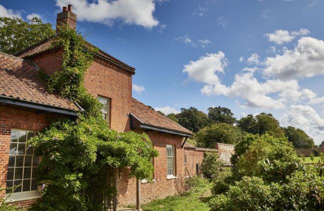 A wisteria-clad, single-storey brick building with large windows overlooks a garden full of shrubs.
